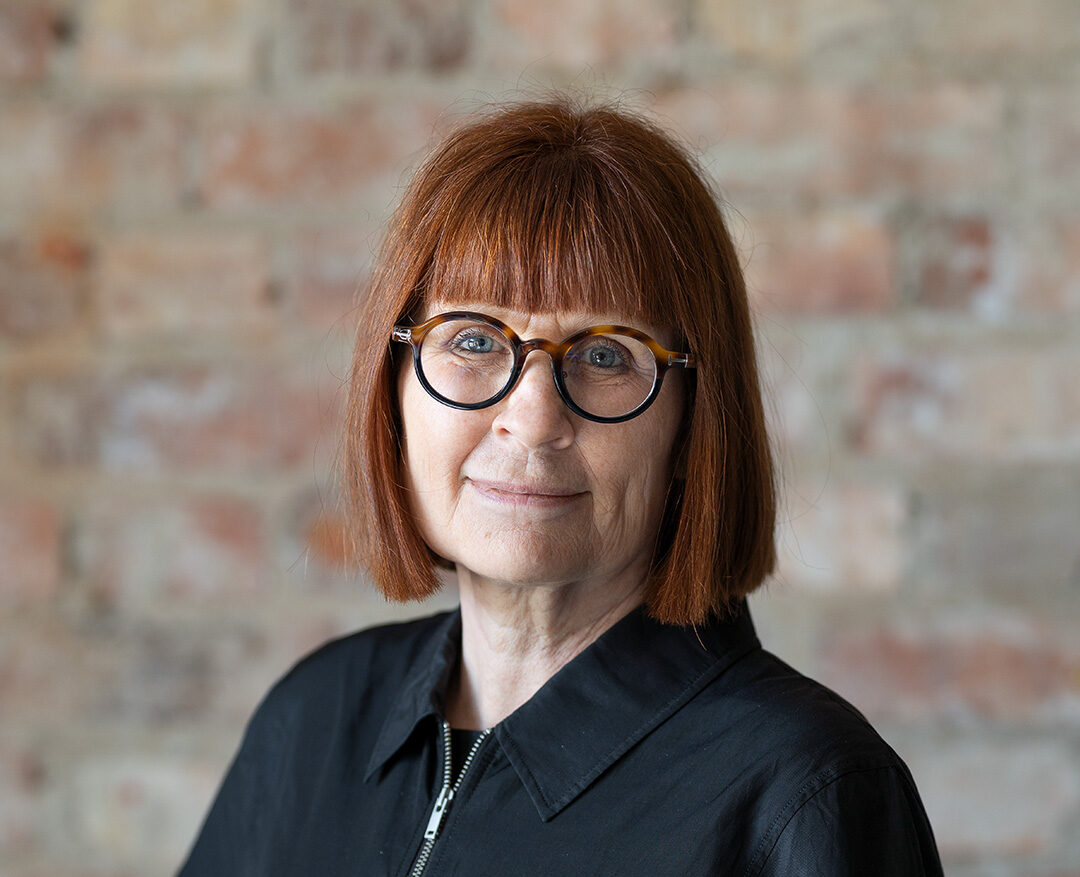 Kvinna med rött hår och glasögon framför en tegelvägg. Styrelse.