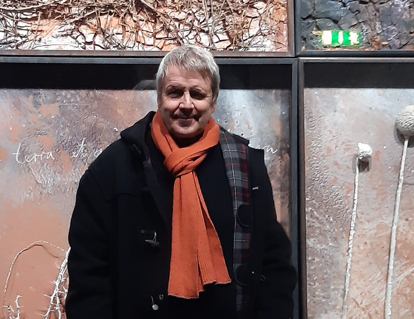 Man i svart jacka och orange halsduk framför konstverk. "terra its