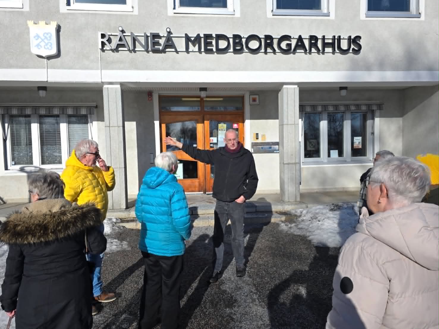 Man pekar mot Råneå Medborgarhus där en grupp besökare lyssnar.